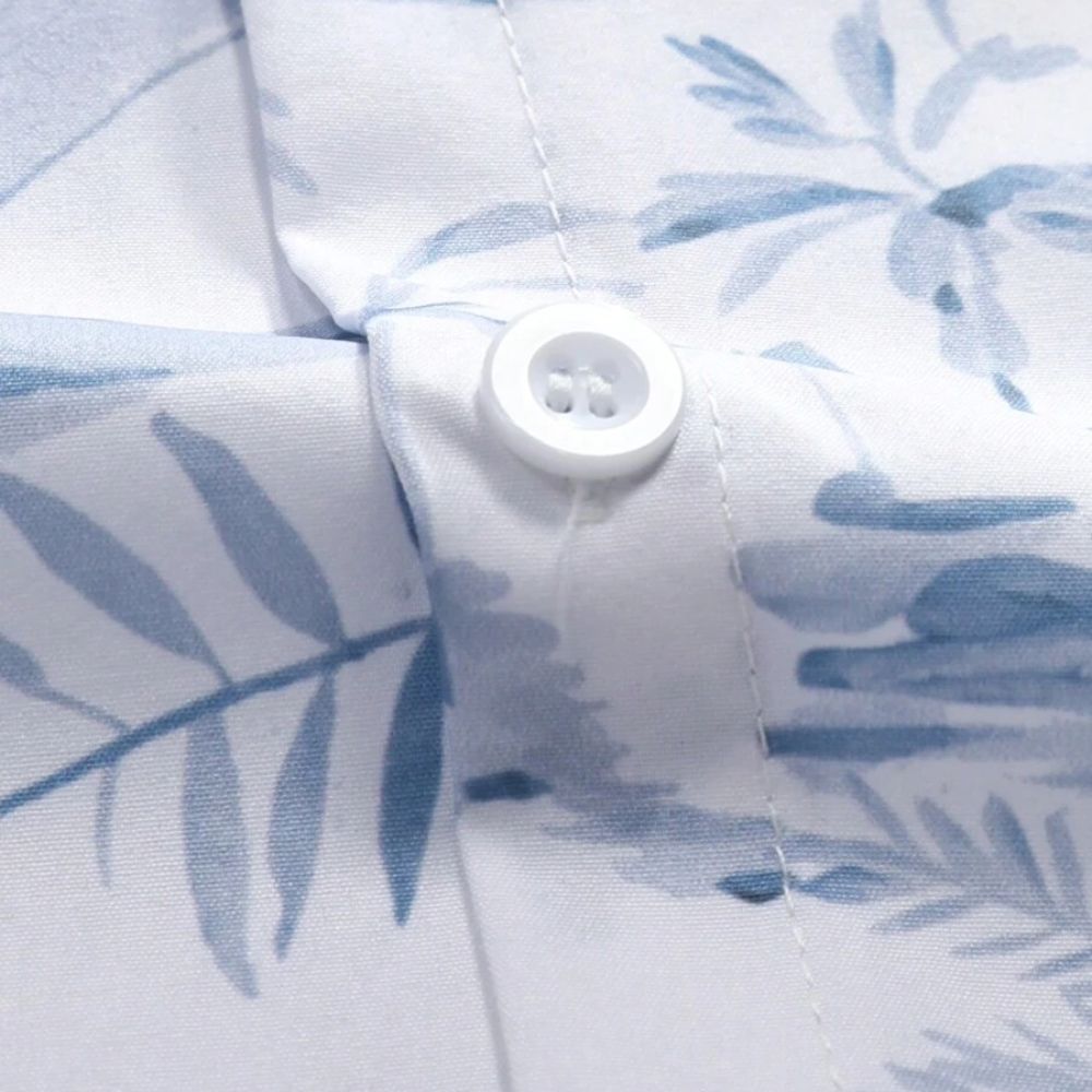 Detalhe do tecido da Camisa Vinser Maragogi, destacando a estampa tropical em tons de azul, o botão branco, as costuras reforçadas e o tecido leve e respirável, evidenciando o acabamento premium da peça