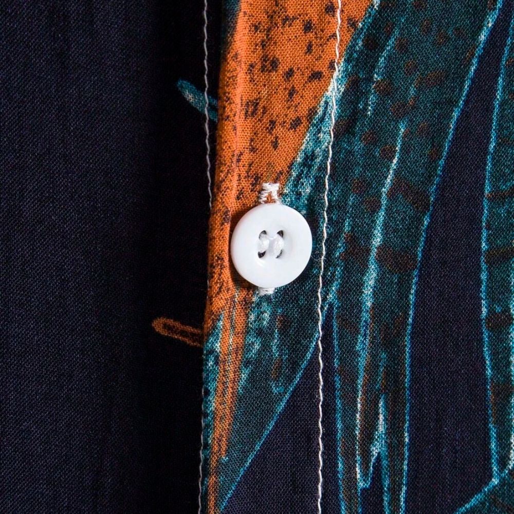 Detalhe do botão da Camisa Vinser Noronha, destacando os botões frontais, as costuras reforçadas, o acabamento refinado e a estampa tropical de folhas em tons de laranja e azul-petróleo sobre fundo azul-marinho, evidenciando a qualidade premium da peça.