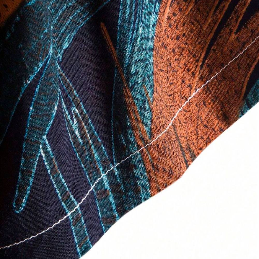Detalhe da costura da Camisa Vinser Noronha, evidenciando o acabamento reforçado, a qualidade da confecção e a estampa tropical de folhas em tons de laranja e azul-petróleo sobre fundo azul-marinho.